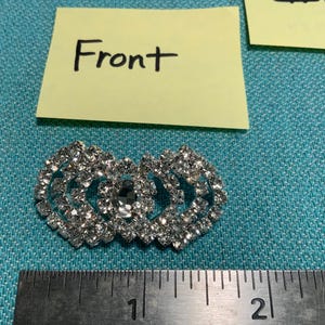 May include: A silver-colored rhinestone brooch with a heart-shaped design. The brooch is made of multiple small rhinestones and has a central, larger rhinestone. The word "Front" is written on a yellow sticky note above the brooch.