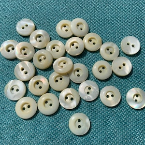 May include: A collection of round, off-white buttons with two holes each, scattered on a textured teal surface. The buttons have a pearlescent sheen, reflecting light. These sewing notions are ideal for various craft and apparel projects.