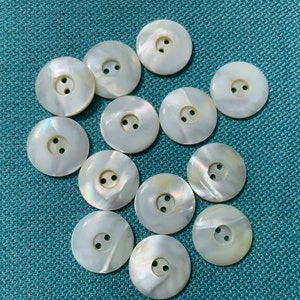 May include: A collection of round, pearlescent white buttons with two holes each. The buttons have a subtle iridescent sheen, and are arranged on a teal-colored surface. These buttons are suitable for sewing and craft projects.