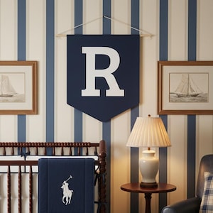 Puede incluir: Un cuarto de niños con papel pintado a rayas azules y blancas, una cuna de madera y arte de veleros enmarcados. Un estandarte azul marino con una "R" blanca cuelga sobre la cuna. Una manta azul marino con un logotipo blanco está sobre la cuna.