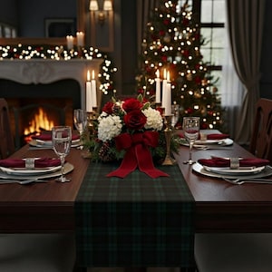May include: A dark green and black plaid table runner centered on a wooden dining table. The runner features a floral centerpiece with red roses, white hydrangeas, and a large red velvet bow, flanked by tall white taper candles in gold holders.