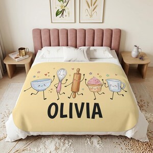 Könnte beinhalten: Eine gelbe Decke mit einer Cartoon-Illustration einer blauen Schüssel, einem Schneebesen, einem hölzernen Nudelholz, einem Cupcake und einem Messbecher, alle mit lächelnden Gesichtern. Der Name OLIVIA steht in großen schwarzen Buchstaben darunter.