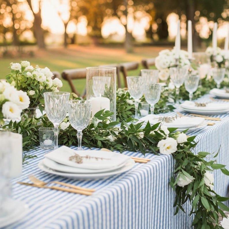 May include: A beautifully set outdoor dining table with a blue and white striped tablecloth. The table is adorned with white plates, crystal wine glasses, gold flatware, and floral arrangements with white flowers and greenery. Tall white candles add to the ambiance.