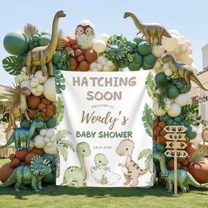 May include: A dinosaur-themed baby shower decoration with a white banner that reads "Hatching Soon Welcome to Wendy's Baby Shower 08.01.2030". The arch is decorated with green, brown, and white balloons, and dinosaur figurines.
