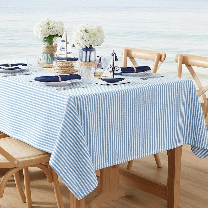 May include: A nautical-themed table setting with a blue and white striped tablecloth. The table is set with white plates, blue napkins, and decorative sailboat figurines. White flowers in glass vases and wooden chairs complete the seaside aesthetic.