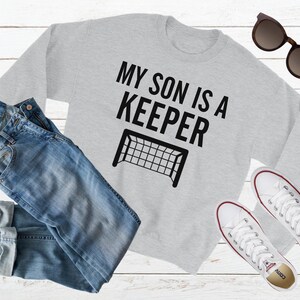 Puede incluir: Sudadera gris con el texto "Mi hijo es un portero" y una silueta negra de una porter&iacute;a de f&uacute;tbol.