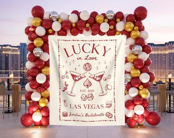 May include: A rectangular white fabric backdrop featuring red line art illustrations of martini glasses, dice, a disco ball, and the text Lucky in Love, EST. 2029, LAS VEGAS, and Jordan's Bachelorette, framed by a large balloon arch of red, white, and gold.