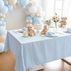 May include: A table set for a celebration, covered with a blue and white striped tablecloth. Teddy bears, a cake, and floral arrangements decorate the table. Balloons in shades of blue and beige form an arch in the background.