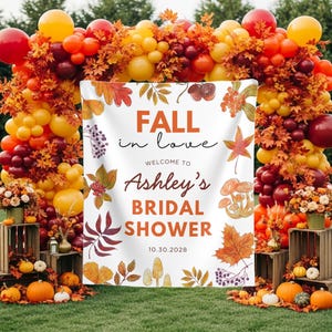 May include: A white rectangular backdrop featuring the text FALL in love WELCOME TO Ashley's BRIDAL SHOWER 10.30.2028, decorated with illustrations of autumn leaves, mushrooms, and berries, framed by a large balloon arch in red, orange, and yellow.