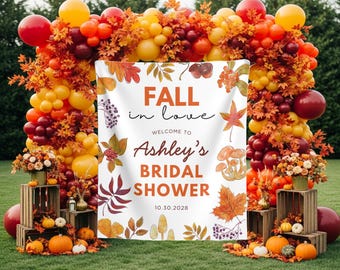 May include: A white rectangular backdrop featuring the text FALL in love WELCOME TO Ashley's BRIDAL SHOWER 10.30.2028, decorated with illustrations of autumn leaves, mushrooms, and berries, framed by a large balloon arch in red, orange, and yellow.