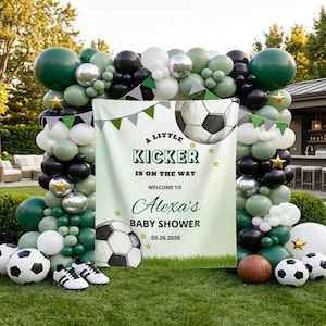 以下が含まれることがあります： A LITTLE KICKER IS ON THE WAY WELCOME TO Alexa's BABY SHOWER 03.26.2030というテキストが書かれたサッカーテーマの長方形のバナー。ダークグリーン、セージ、白、黒、銀のバルーンアーチとサッカーボールで装飾されています。
