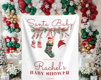 May include: A white banner with the text "Santa Baby" and "Rachel's Baby Shower" in red script. The banner features Christmas decorations, including a stocking, mittens, and a Santa hat. The background includes Christmas trees, gifts, and a balloon arch.