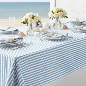 May include: A coastal-themed table setting with a blue and white striped tablecloth. The table is set with white plates, blue napkins, champagne flutes, and starfish accents. White roses in glass vases and seashells complete the beachy aesthetic.