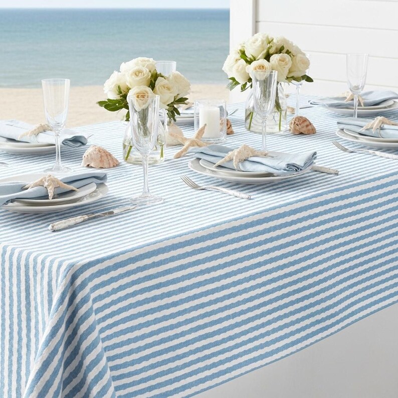 May include: A coastal-themed table setting with a blue and white striped tablecloth. The table is set with white plates, blue napkins, champagne flutes, and starfish accents. White roses in glass vases and seashells complete the beachy aesthetic.