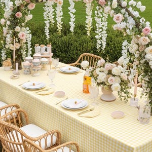 Pode incluir: Uma mesa de jantar ao ar livre lindamente montada com uma toalha de mesa xadrez amarela e branca. A mesa é adornada com pratos brancos, talheres dourados e centros de mesa florais. Cadeiras de vime com almofadas brancas são dispostas ao redor da mesa.