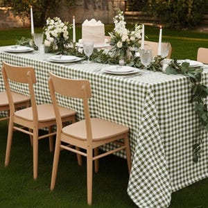 May include: A beautifully set outdoor dining table with a green and white checkered tablecloth. The table is adorned with white flowers, candles, and a cake. Light-colored wooden chairs are arranged around the table, creating an inviting atmosphere.