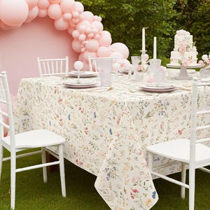May include: A beautifully set table for a party, featuring a floral tablecloth, white chairs, and pink accents. The table is set with plates, glasses, and silverware. A cake and candles add to the festive atmosphere. A pink balloon arch is in the background.