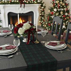 May include: A dark green and black plaid table runner centered on a dark wooden dining table. The table is set with white plates, maroon cloth napkins in silver rings, clear wine glasses, and a centerpiece of red roses and white hydrangeas.