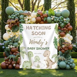 May include: A baby shower decoration featuring a banner with the text "Hatching Soon" and "Welcome to Wendy's Baby Shower." The banner is surrounded by a balloon arch in shades of green, brown, and cream, with dinosaur figurines.