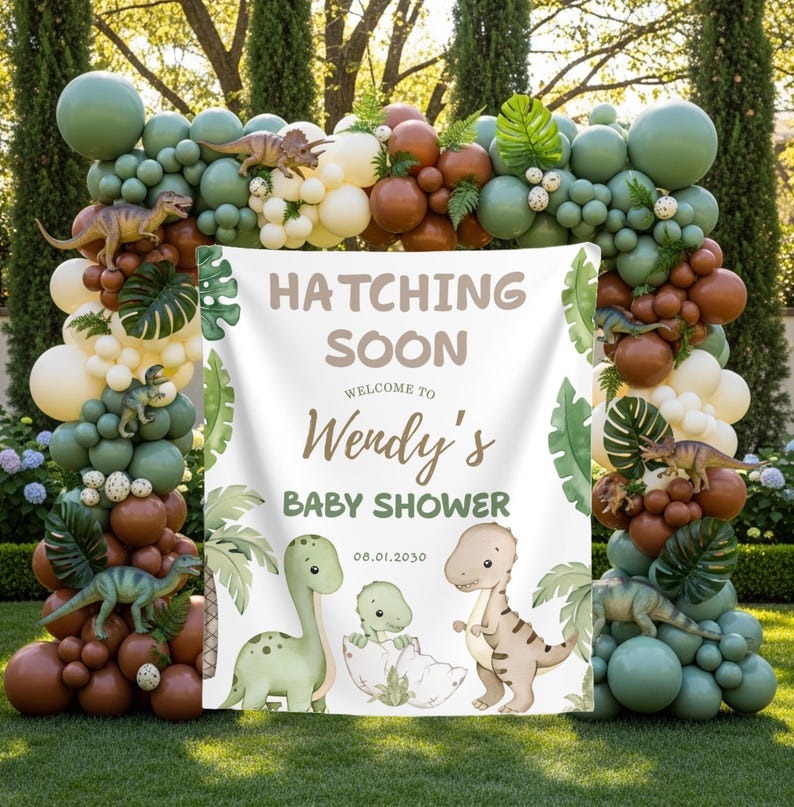 May include: A baby shower decoration featuring a banner with the text "Hatching Soon" and "Welcome to Wendy's Baby Shower." The banner is surrounded by a balloon arch in shades of green, brown, and cream, with dinosaur figurines.