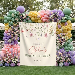 Pu&ograve; includere: Un banner per un addio al nubilato con dettagli floreali e la scritta "Welcome to Chloe's Bridal Shower 07.25.2027." Il banner &egrave; color crema ed &egrave; incorniciato da un arco di palloncini colorati e composizioni floreali.