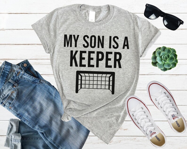 Puede incluir: Camiseta gris con texto negro que dice "Mi hijo es un portero" y una silueta negra de una porter&iacute;a de f&uacute;tbol.