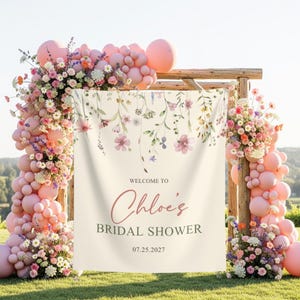 May include: A floral archway with pink balloons and flowers frames a banner that reads "Welcome to Chloe's Bridal Shower 07.25.2027". The arch is made of wood and set on a grassy lawn.