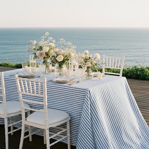 May include: A beautifully set table with a blue and white striped tablecloth, white chairs, and floral centerpieces. The table is set with plates, glasses, and silverware. The ocean is visible in the background.