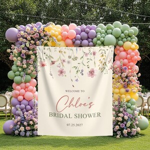 Pu&ograve; includere: Un cartello decorativo per un addio al nubilato, con la scritta "Welcome to Chloe's Bridal Shower" e la data 07.25.2027. Il cartello &egrave; incorniciato da un arco colorato di palloncini e composizioni floreali nei toni del rosa, viola, giallo e verde.