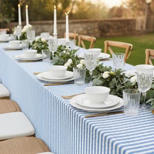 Puede incluir: Una mesa bellamente decorada con un mantel a rayas azules y blancas. Platos, cuencos y copas de cristal blancos están dispuestos, complementados con cubiertos dorados y velas encendidas. Los arreglos florales añaden un toque de elegancia al comedor al aire libre.