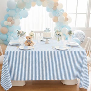 May include: A table set for a celebration, featuring a blue and white striped tablecloth. The table is decorated with a cake, flowers, and a teddy bear. A balloon arch in shades of blue and white frames the scene.