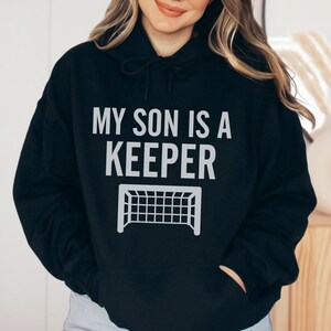 Puede incluir: Sudadera con capucha negra con un gr&aacute;fico blanco que dice "Mi hijo es un portero" con una porter&iacute;a de f&uacute;tbol debajo del texto.