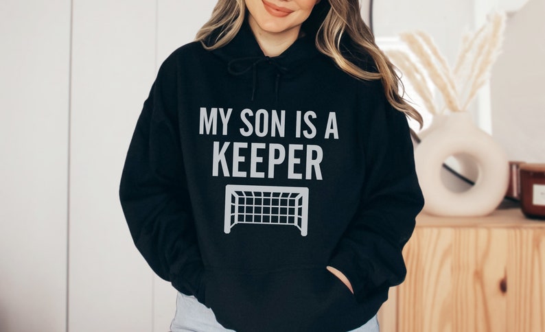Puede incluir: Sudadera con capucha negra con un gr&aacute;fico blanco que dice "Mi hijo es un portero" con una porter&iacute;a de f&uacute;tbol debajo del texto.