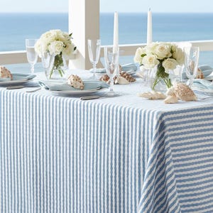 May include: A dining table set for a seaside theme. The table is covered with a blue and white striped tablecloth. White roses, seashells, and candles decorate the table. Clear glasses and plates are set for each place setting.