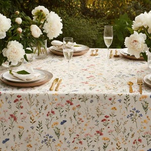 May include: A beautifully set table with a floral tablecloth featuring a pattern of colorful wildflowers. The table is set with plates, gold cutlery, champagne flutes, and white napkins. White peony flowers in vases add a touch of elegance.