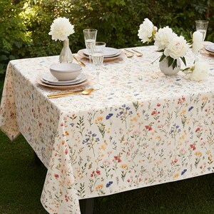 May include: A rectangular tablecloth with a floral pattern in various colors, including red, blue, and yellow, covers a table set for a meal. White flowers in vases and glasses of liquid are on the table. Gold-colored utensils and plates are also visible.