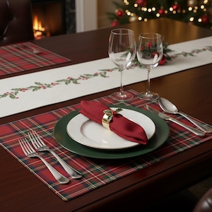 Puede incluir: Una mesa festiva con un mantel individual a cuadros rojos y verdes, un plato blanco con borde dorado y un plato verde. Una servilleta roja está sujeta por un anillo dorado con detalles de acebo. Copas de vino y cubiertos presentes.