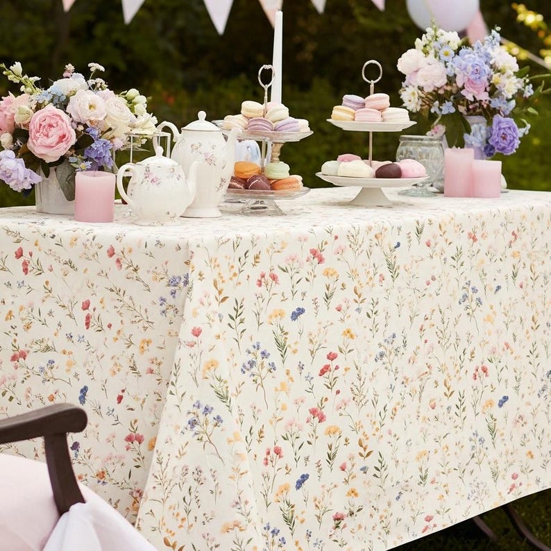 May include: A table set for a garden party, featuring a floral tablecloth with a pattern of small, colorful wildflowers. The table is adorned with bouquets of pink and purple flowers, teacups, teapots, and tiered serving platters of macarons and pastries.