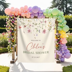 Pu&ograve; includere: Un arco in legno decorato con una ghirlanda di palloncini pastello e composizioni floreali. Uno striscione color crema mostra il testo "Welcome to Chloe's Bridal Shower" e la data 07.25.2027.
