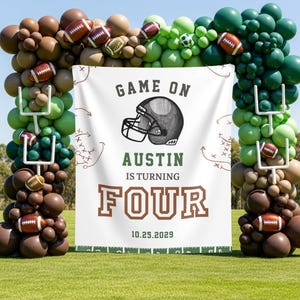 May include: A football-themed party decoration featuring a balloon arch in shades of brown and green, adorned with footballs and goalpost accents. A banner displays the text "Game On" and "Austin is turning FOUR" with the date 10.25.2029.