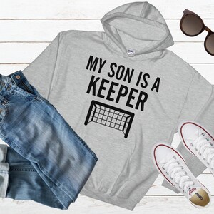 Puede incluir: Sudadera con capucha gris con texto negro que dice "Mi hijo es un portero" y una silueta negra de una porter&iacute;a de f&uacute;tbol.