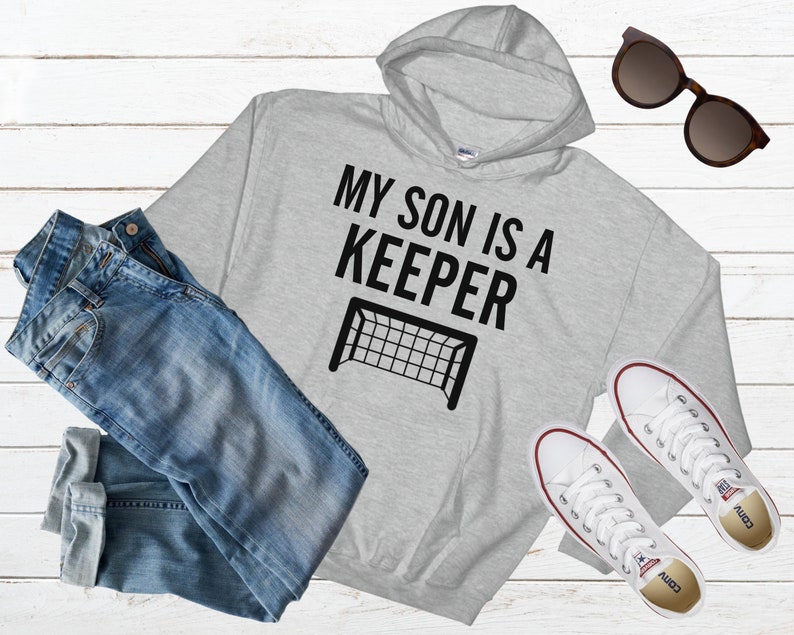 Puede incluir: Sudadera con capucha gris con texto negro que dice "Mi hijo es un portero" y una silueta negra de una porter&iacute;a de f&uacute;tbol.