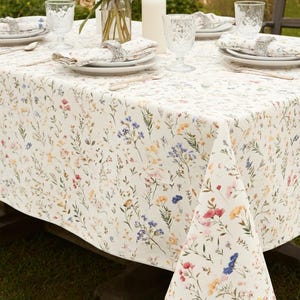 May include: A cream-colored tablecloth with a delicate floral pattern in shades of pink, blue, yellow, and green. The table is set with white plates, napkins, and clear glasses, creating an elegant dining scene. Silver napkin rings add a touch of sophistication.