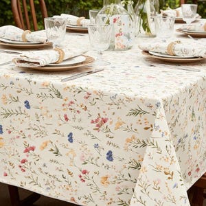 May include: A dining table set for a meal, featuring a cream-colored tablecloth with a floral pattern in pink, yellow, and blue. The table is set with plates, napkins, glasses, and cutlery, creating an inviting atmosphere for a gathering.
