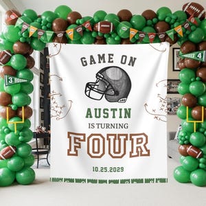 May include: A football-themed party decoration featuring a banner with the text "Game On, Austin is turning Four" and a football helmet graphic. The banner is surrounded by green and brown balloons, footballs, and pennants.