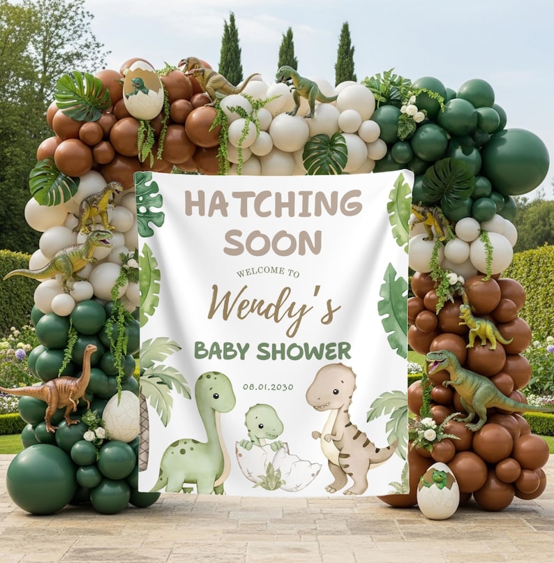 May include: A baby shower decoration featuring a banner with the text "Hatching Soon" and "Welcome to Wendy's Baby Shower". The banner is surrounded by a balloon arch in shades of brown, green, and white, with dinosaur figurines and tropical leaves.