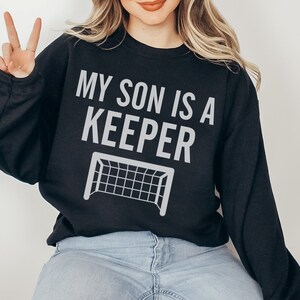 Puede incluir: Sudadera negra con el texto "MY SON IS A KEEPER" y una silueta blanca de una porter&iacute;a de f&uacute;tbol.