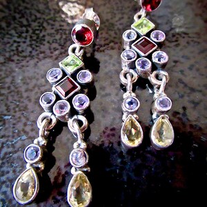 May include: A pair of silver dangle earrings with multiple gemstones. The earrings feature a teardrop-shaped bottom with a green, red, and purple gemstone cluster.