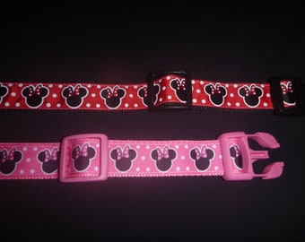May include: Two dog collars with a repeating Minnie Mouse head pattern. One is red with white polka dots and black Minnie Mouse heads. The other is pink with a repeating Minnie Mouse head pattern. Both have plastic buckles.