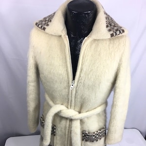 K&ouml;nnte beinhalten: Ein wei&szlig;er Strickcardigan mit braunem und wei&szlig;em Muster an Kragen und Taschen. Der Cardigan hat einen Rei&szlig;verschluss und einen G&uuml;rtel.